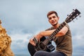 Young man with acoustic guitar sitting on beach surrounded with rocks on rainy day using smartphone Royalty Free Stock Photo