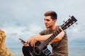 Young man with acoustic guitar sitting on beach surrounded with rocks on rainy day using smartphone Royalty Free Stock Photo