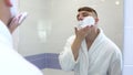 A young man applies shaving foam in front of a mirror. A man in a white coat with foam on his face. View through the Royalty Free Stock Photo