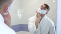 A young man applies shaving foam in front of a mirror. A man in a white coat with foam on his face. View through the Royalty Free Stock Photo