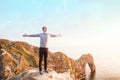 Young man arms raised up to sky, celebrating freedom standing on the rock with Durdle Door view at sunset. Positive emotions feeli