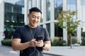 A young man, an Asian sportsman, is standing on a city street in headphones, holding a phone in his hands, making a call Royalty Free Stock Photo