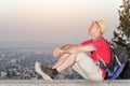 Young man in backpack and hat sitting on background of large city below. Side view