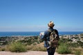 A young man with a backpack and a map of the area in his hands stands on a mountain near the city and the sea Royalty Free Stock Photo