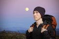 Young man with backpack in mountains. Hilly night landscape with the moon. Royalty Free Stock Photo