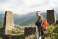 Young man with backpack standing on stone near old towers on background of mighty mountains. Male tourist enjoying beautiful view