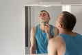 Young man in bathroom removes his beard with razor, shaving foam on face. Face care. Reflection in mirror Royalty Free Stock Photo