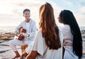 A young man at the beach playing his guitar with his two female friends. Diverse group of friends listening music on a Royalty Free Stock Photo