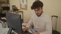 Young man with beard and glasses explores smartphone while working in a modern office space equipped with computer and suitcase, Royalty Free Stock Photo