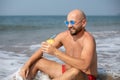 Vacationer in sunglasses holding drink decorated with pineapple and smiling sitting in wave on shore on sunny day. Royalty Free Stock Photo