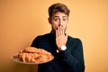 Young man with beard holding plate with croissants standing over isolated yellow background cover mouth with hand shocked with