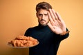 Young man with beard holding plate with croissants standing over isolated yellow background with open hand doing stop sign with