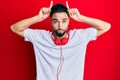 Young man with beard listening to music using headphones doing funny gesture with finger over head as bull horns