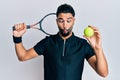 Young man with beard playing tennis holding racket and ball making fish face with mouth and squinting eyes, crazy and comical