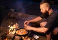 A young man with a beard prepares a dish of bacon with scrambled eggs on a fire. Cooking and outdoor recreation in summer Royalty Free Stock Photo