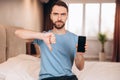 Young man with beard sitting in bed and shows disappoint and does not like using phone after sleep while. Royalty Free Stock Photo