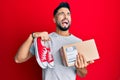 Young man with beard taking casual red shoes from box angry and mad screaming frustrated and furious, shouting with anger looking