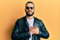 Young man with beard wearing black leather jacket and sunglasses smiling with hands on chest with closed eyes and grateful gesture Royalty Free Stock Photo