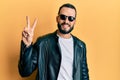 Young man with beard wearing black leather jacket and sunglasses smiling with happy face winking at the camera doing victory sign Royalty Free Stock Photo