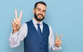 Young man with beard wearing business vest smiling looking to the camera showing fingers doing victory sign Royalty Free Stock Photo