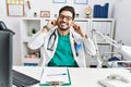 Young man with beard wearing doctor uniform and stethoscope at the clinic smiling pulling ears with fingers, funny gesture