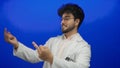 Young man with a beard wearing a white coat humorously gestures with middle fingers against a solid blue background, smiling and Royalty Free Stock Photo