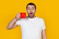 Young man with a beard in a white t-shirt drinks a cup of coffee, scared in shock with a surprised face, scared and excited with