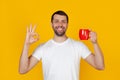 Young man with a beard in a white T-shirt smiling, holding a red cup of coffee and showing the gesture, everything is okay, a man Royalty Free Stock Photo