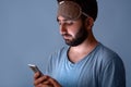 Young man with black beard standing isolated over blue background, having sleeping mask on his forehead, holding his smartphone in Royalty Free Stock Photo