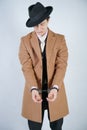 Young man in a black business suit and beige fashion coat with a stylish hat stands in police handcuffs on a white solid Studio ba Royalty Free Stock Photo