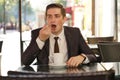 A young man in a black business suit, white shirt and tie sits in a city street cafe at a table and enjoys his cappuccino with foa Royalty Free Stock Photo