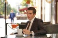A young man in a black business suit, white shirt and tie is sitting in a city street cafe at a table and enjoying a Cup of coffee Royalty Free Stock Photo