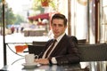 A young man in a black business suit, white shirt and tie is sitting in a city street cafe at a table and enjoying a Cup of coffee Royalty Free Stock Photo