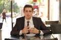 A young man in a black business suit, white shirt and tie sits in a city street cafe at a table and enjoys his cappuccino with foa Royalty Free Stock Photo