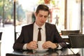 a young man in a black business suit, white shirt and tie sits in a city street cafe at a table and enjoys his cappuccino with foa Royalty Free Stock Photo