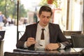 A young man in a black business suit, white shirt and tie sits in a city street cafe at a table and enjoys his cappuccino with foa Royalty Free Stock Photo