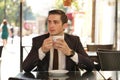 A young man in a black business suit, white shirt and tie sits in a city street cafe at a table and enjoys his cappuccino with foa Royalty Free Stock Photo