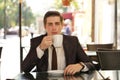 A young man in a black business suit, white shirt and tie sits in a city street cafe at a table and enjoys his cappuccino with foa Royalty Free Stock Photo