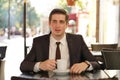 A young man in a black business suit, white shirt and tie sits in a city street cafe at a table and enjoys his cappuccino with foa Royalty Free Stock Photo