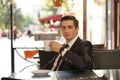 A young man in a black business suit, white shirt and tie sits in a city street cafe at a table and enjoys his cappuccino with foa Royalty Free Stock Photo