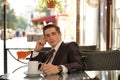 A young man in a black business suit, white shirt and tie is sitting in a city street cafe at a table and enjoying a Cup of coffee Royalty Free Stock Photo