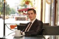 A young man in a black business suit, white shirt and tie is sitting in a city street cafe at a table and enjoying a Cup of coffee Royalty Free Stock Photo