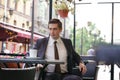 A young man in a black business suit, white shirt and tie is sitting in a city street cafe at a table and enjoying a Cup of coffee Royalty Free Stock Photo