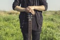 A young man in black clothes is holding a guitar with both hands against the blue sky and green grass Royalty Free Stock Photo