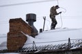 A young man in black dresses is standing on a red roof and cleaning the chimney with a metal brush on a long cable.