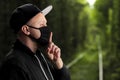 Young man in black protective antiviral mask with a finger to his lips in the summer park. The guy is resting outdoors in a mask Royalty Free Stock Photo