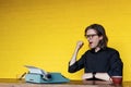 Portrait of a man in black shirt glass, showing successful, seated at a table near typewriter over yellow background. Royalty Free Stock Photo