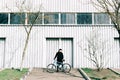 A young man in black sportswear holds a Bicycle standing next to him, against the background of a light hangar on a Sunny spring Royalty Free Stock Photo