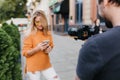 Young man in black t-shirt making photo of glad blonde girl in orange sweater. Attractive woman in glasses holding