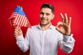 Young man with blue eyes holding flag of united states of america over red isolated background doing ok sign with fingers, Royalty Free Stock Photo